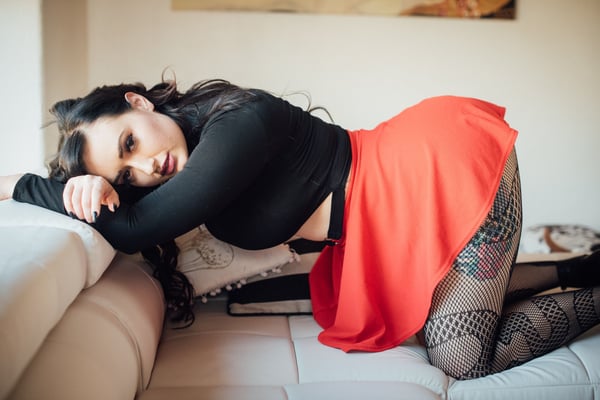 A woman with long dark hair poses reclined on a couch in a black top, red skirt, and patterned stockings, with tattoos visible on her leg.