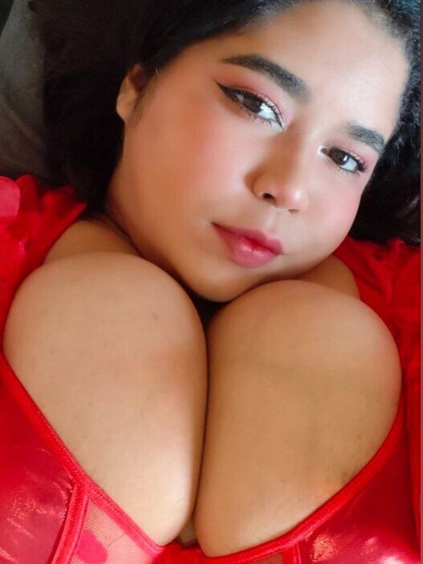 Close-up portrait of a woman with dark hair and bold makeup, wearing a red lace top, lying down indoors.