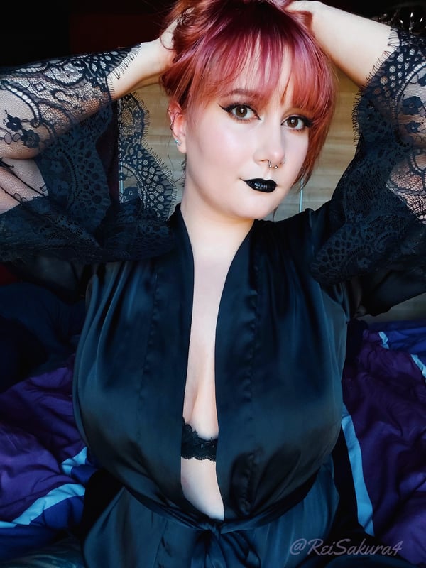 A woman with dyed red hair and dark lipstick, wearing a black lace robe, posing indoors with a confident expression.