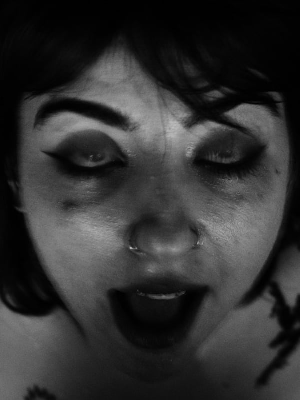 Close-up portrait of a person with dark hair, dramatic makeup, and a nose piercing, making an expressive open-mouthed face in black and white.