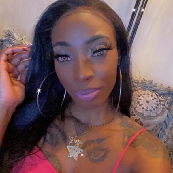 A woman with long dark hair, large hoop earrings, and a star pendant necklace poses confidently indoors, wearing a pink top.