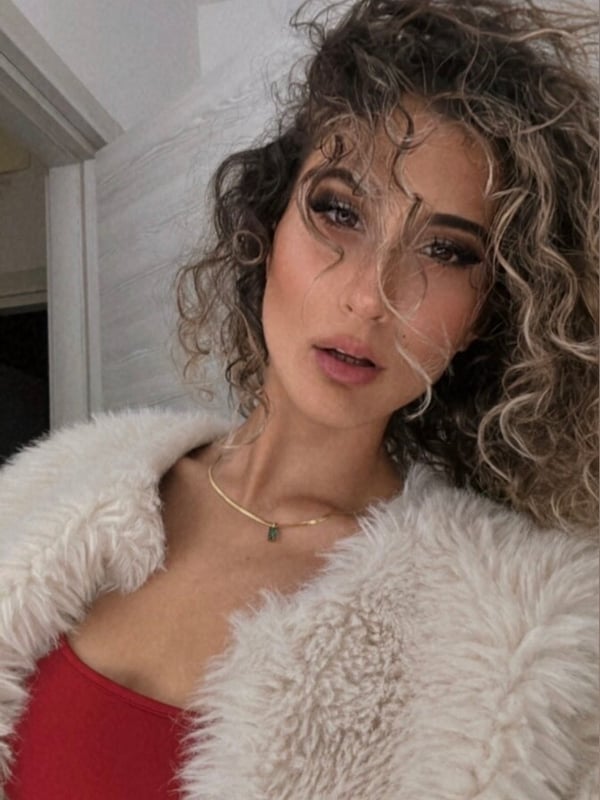 A woman with curly blonde hair wearing a red top and white fur jacket, posing with a sultry expression indoors.