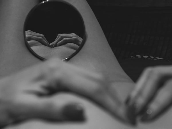 A black and white image of hands holding a round mirror against the body, reflecting a detailed view.