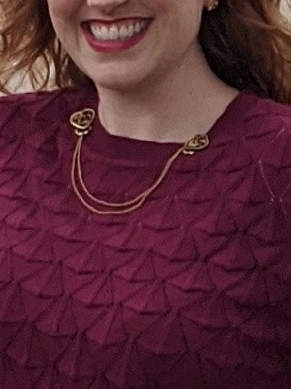 Smiling woman with red curly hair and gold jewelry, wearing a textured maroon sweater, standing on a green path outdoors.