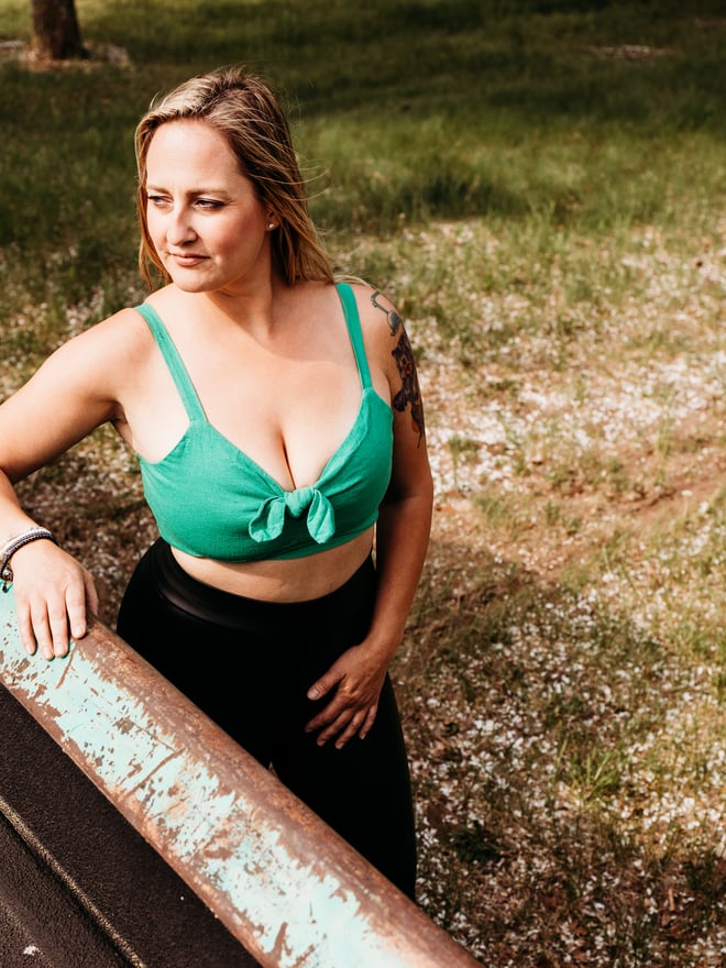 Woman standing outdoors next to a vintage van in a green bikini top and black bottoms, smiling against a grassy background.