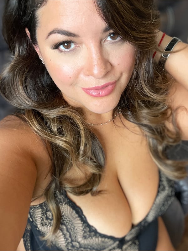 Close-up selfie of a woman with long wavy brown hair, smiling confidently while wearing a lace top, accessorized with a bracelet and necklace.