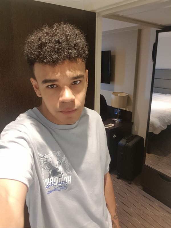 A young man with curly hair wearing a light gray t-shirt, standing in a modern hotel room near a door.
