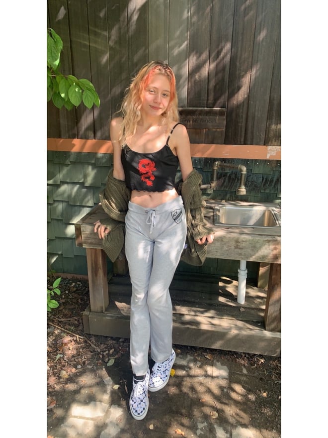 Young woman with long wavy orange-dyed hair wearing a black tank top and light gray jacket, posing casually outdoors near a wooden structure with greenery in the background.