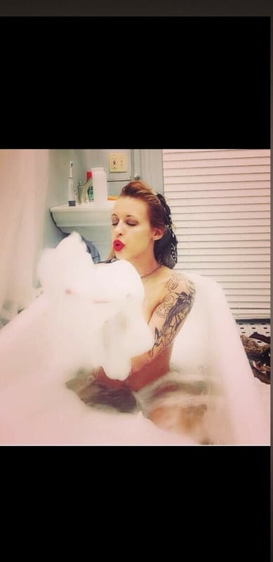 A woman with tattoos lounging in a bubble bath, smiling playfully in a bathroom setting.