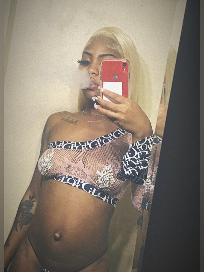 A woman with long blonde hair taking a mirror selfie while smoking, wearing lace lingerie, with tattoos visible on her shoulder.