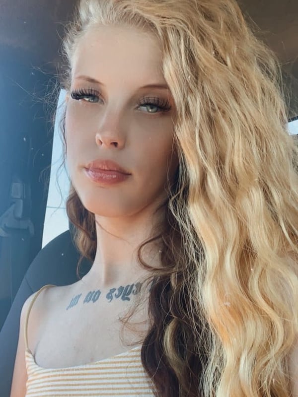 Close-up selfie of a young woman with long blonde wavy hair, wearing a striped tank top, sitting in a car with natural light.