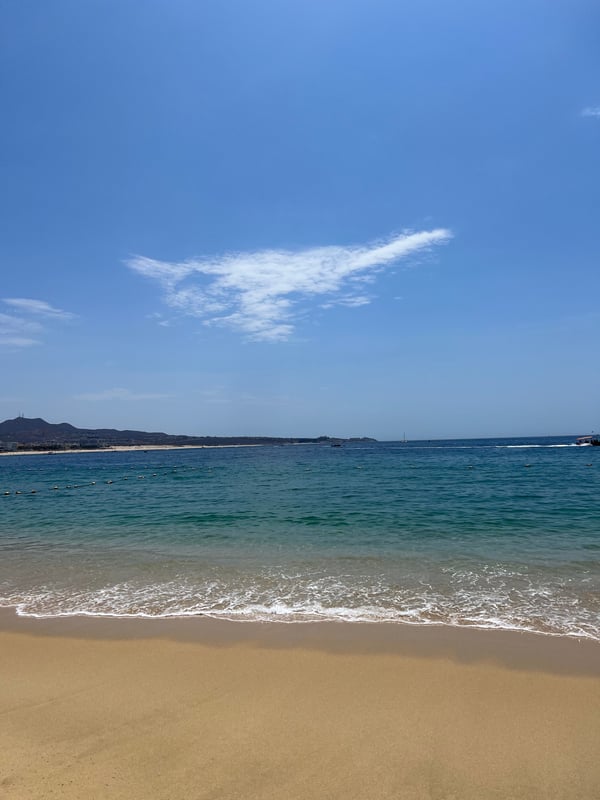 Scenic beach landscape with golden sand, turquoise ocean waves, distant hills, and a clear blue sky with scattered clouds.