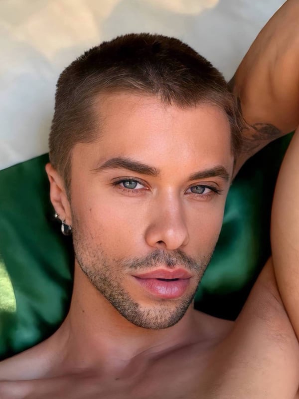 Close-up portrait of a young man with short blonde hair, blue eyes, and piercings, posing shirtless against a green background.