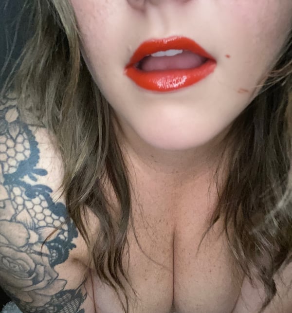 Close-up portrait of a woman with blonde hair, red lipstick, and lace tattoos on her shoulder, posing with an open mouth expression.