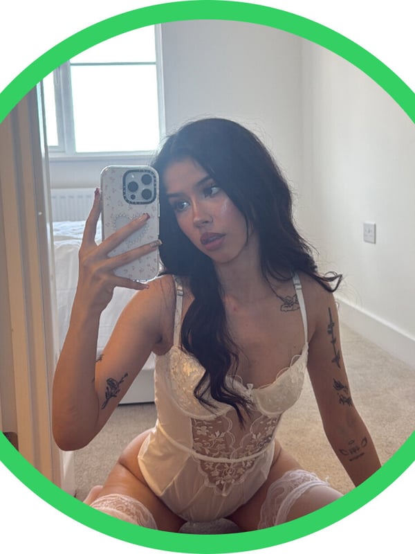 Young woman with long dark hair taking a mirror selfie in a white lace outfit, sitting in a modern bedroom.