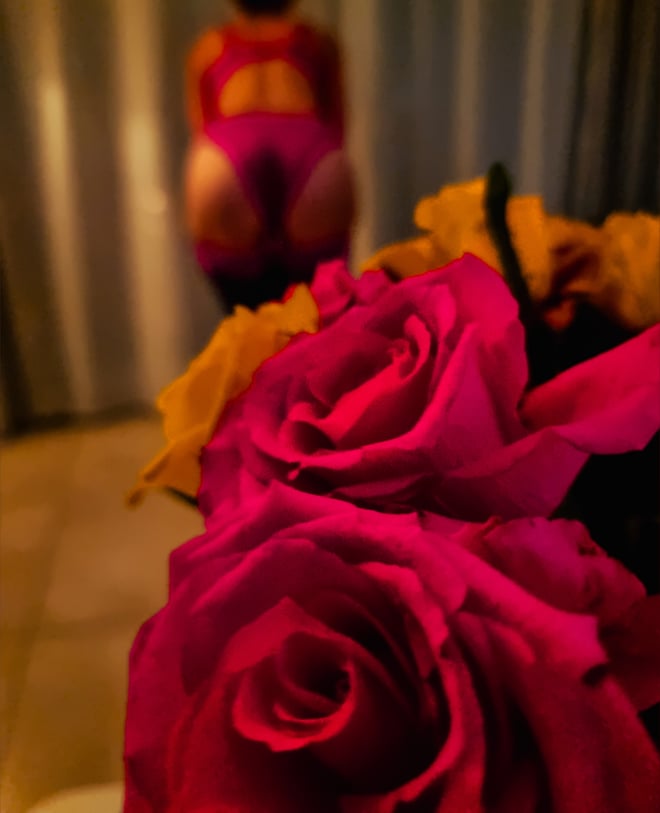 A woman in a pink outfit posing from behind near a vase of colorful roses in a softly lit room.