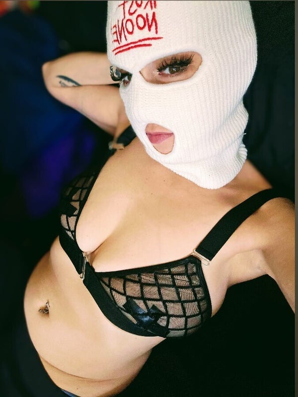 Close-up portrait of a woman wearing a white balaclava mask with red 'MOON' text, lace bralette, and visible tattoos and piercing, posing confidently.