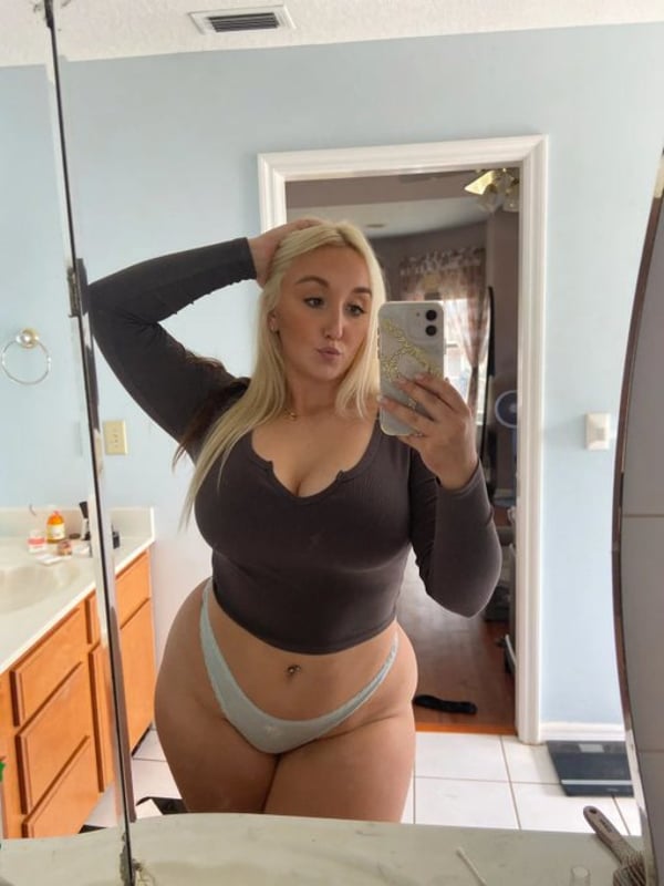A smiling blonde woman taking a selfie in a white bikini top indoors.