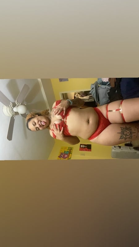 A woman standing in a room with a ceiling fan, wearing red lingerie and posing confidently.