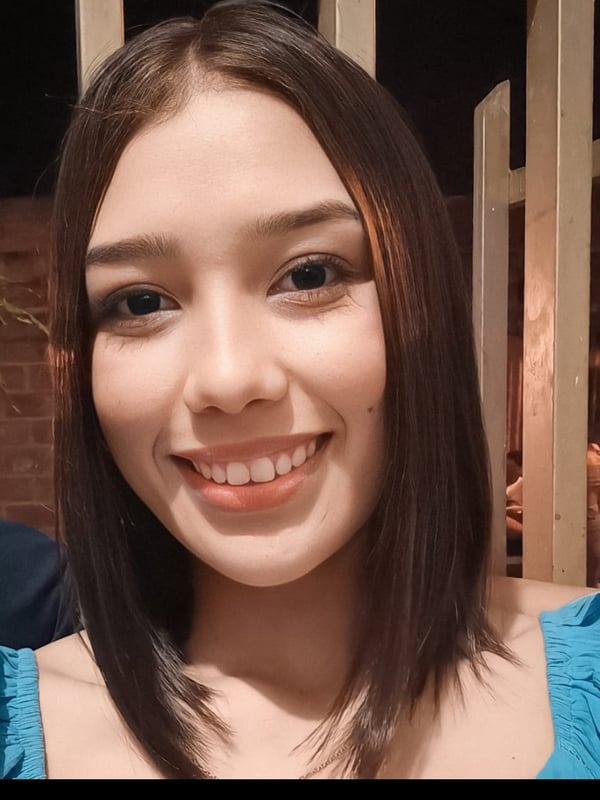 A young woman with shoulder-length brown hair smiles warmly at the camera outdoors at night, wearing a blue off-shoulder top.