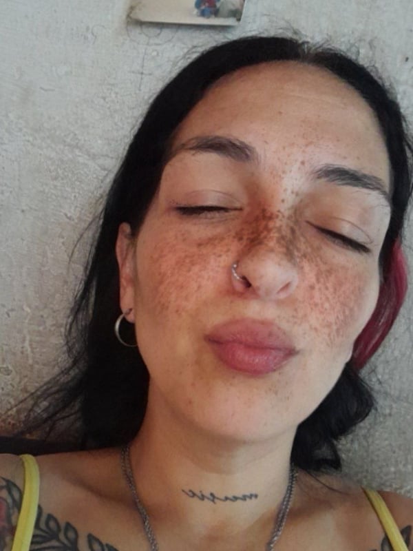 Close-up portrait of a woman with freckles, dark hair, piercings, and tattoos, making a playful duck face while lying down.