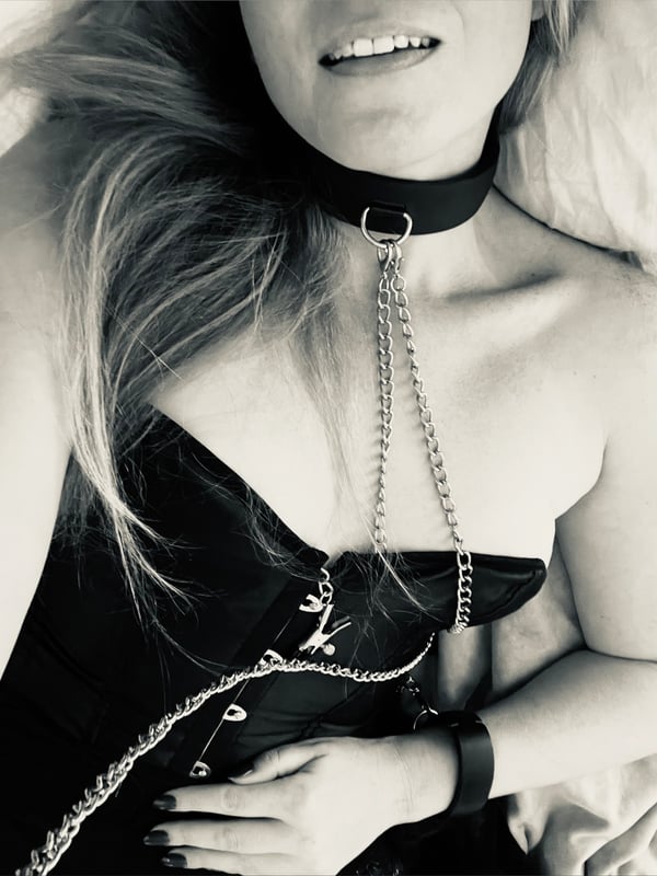 A woman with long blonde hair smiling, wearing a choker necklace with chains, lying down in a black and white photo.