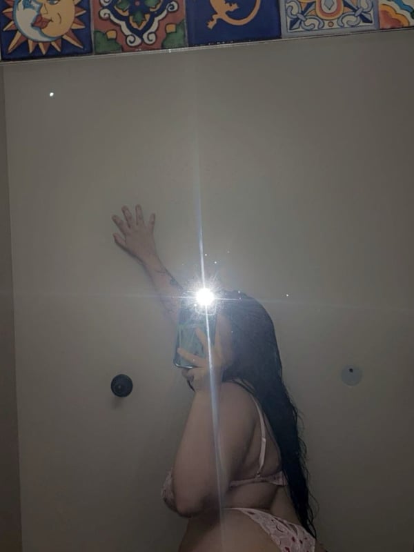 Young woman with long dark hair standing in a pink swimsuit in front of a bathroom mirror with decorative tiled borders.
