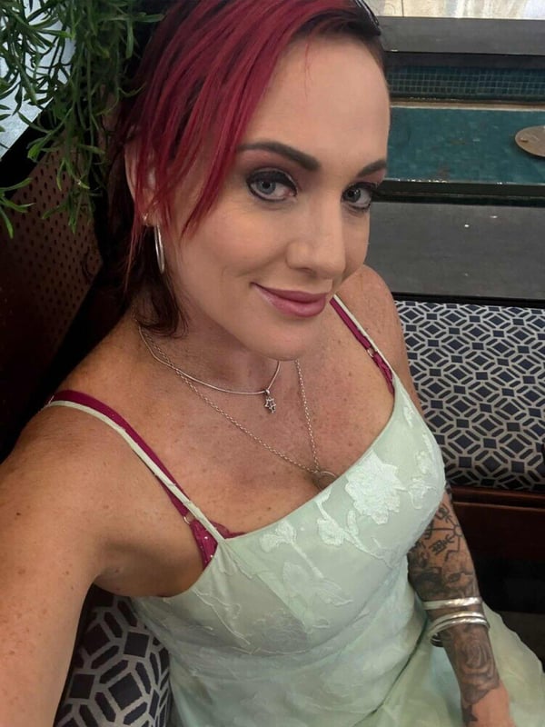 Woman with red hair and tattoos smiling at the camera while seated indoors near a window with plants.