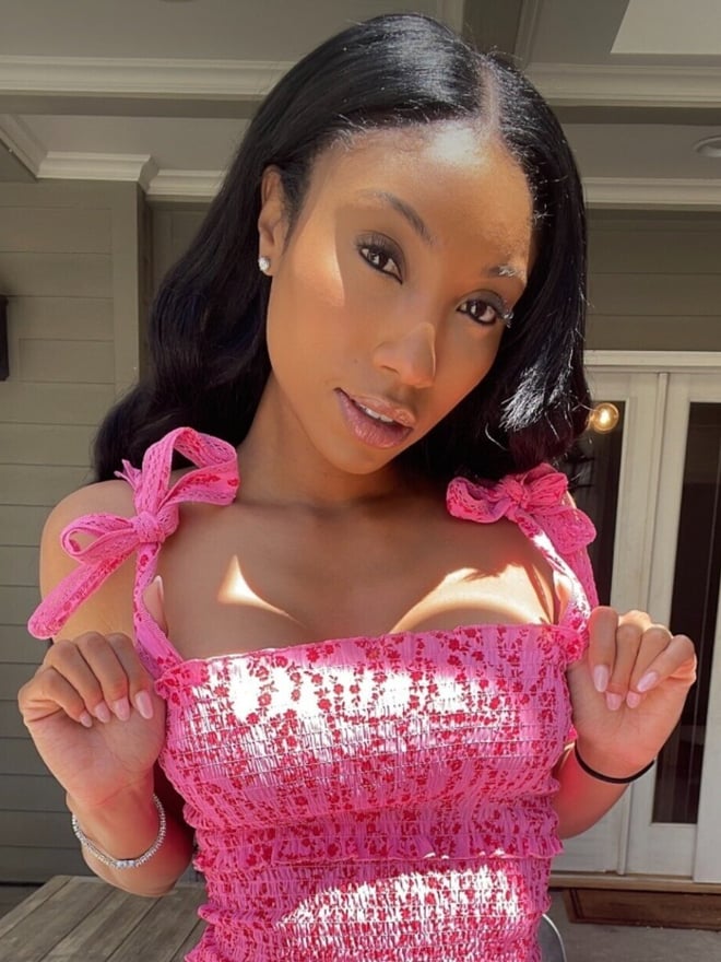 A woman in a pink spaghetti strap top posing on a porch, smiling with sunlight highlighting her features.