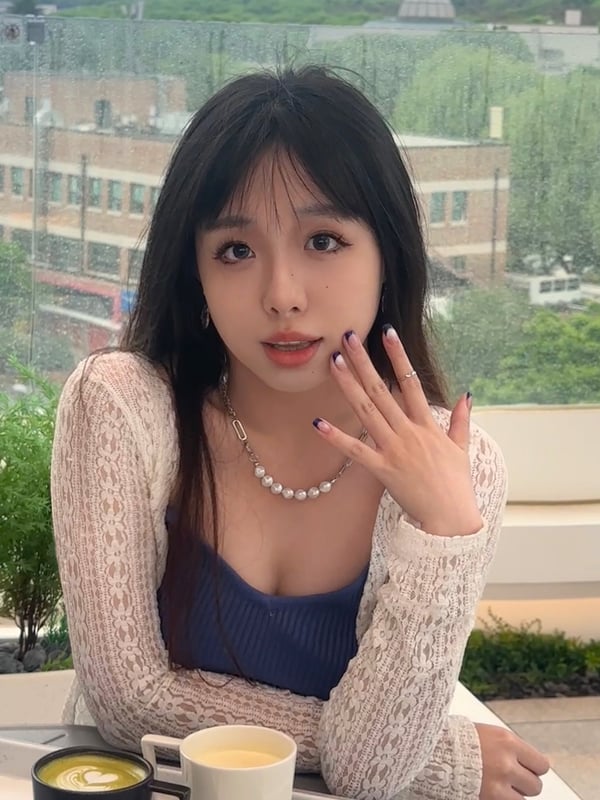 Young woman with straight black hair sitting at an outdoor table, holding a cup of coffee, wearing a lace cardigan over a blue top, with cityscape view in the background.