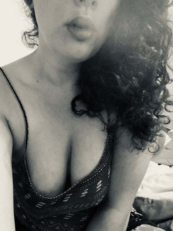 A close-up selfie of a woman with curly hair puckering her lips, wearing a low-cut top.