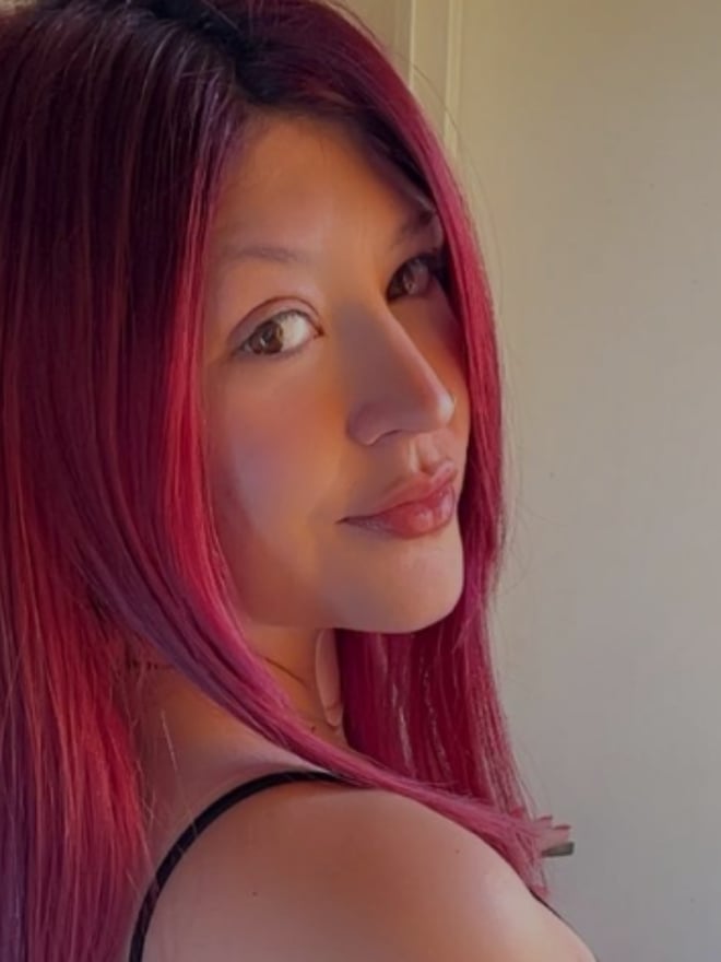 Woman with long pink hair smiling at the camera, wearing a black top, against a light indoor background.