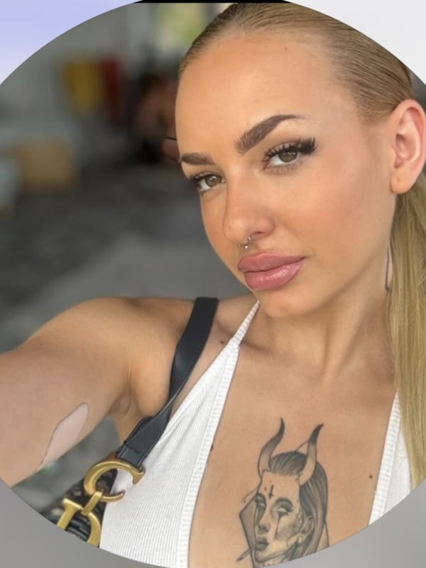 Close-up portrait of a young blonde woman with striking makeup, a tattoo visible on her chest, wearing a white top and gold necklace.