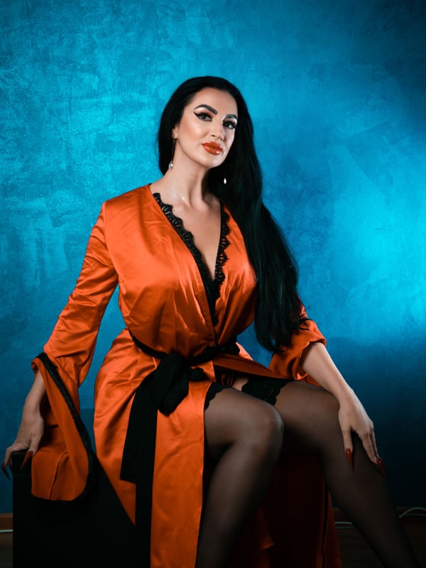 A woman with long dark hair posing elegantly in an open orange satin robe against a blue background.