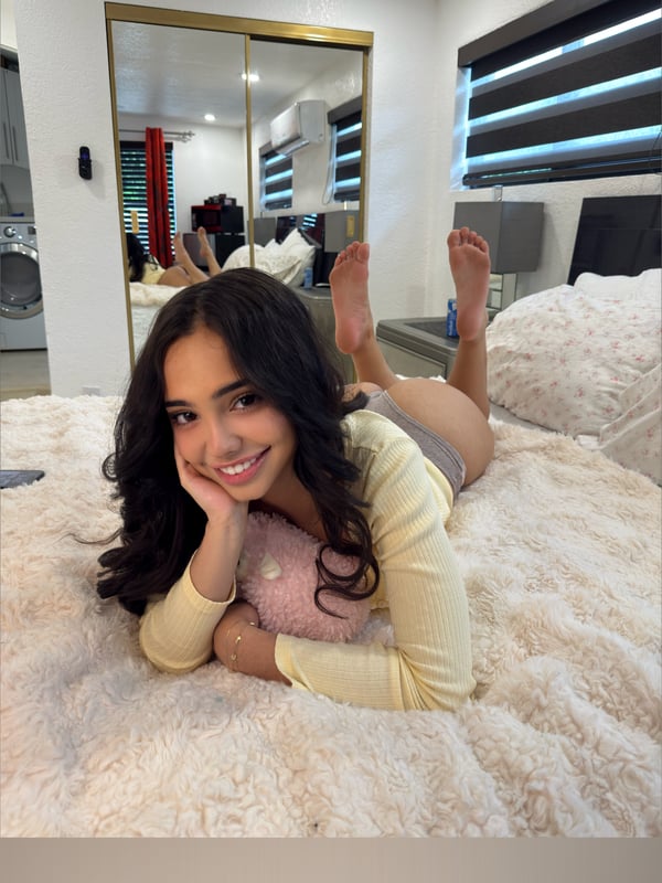 Young woman with long dark hair lying on a bed in a bedroom, smiling at the camera while propped on pillows near a mirror.