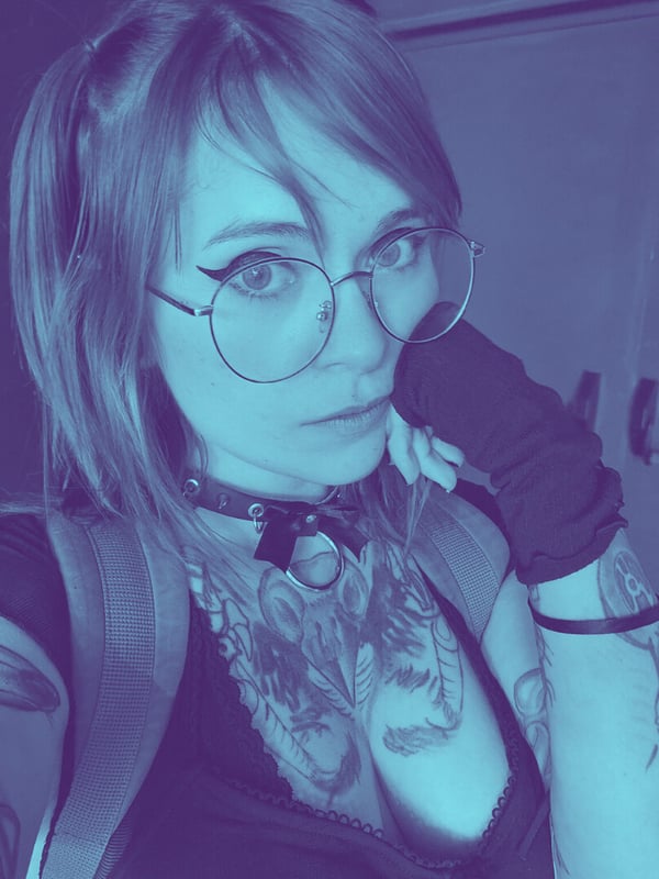 Young woman with red hair, glasses, choker, and visible tattoos, posing confidently indoors in a dark top and gloves with blue lighting.