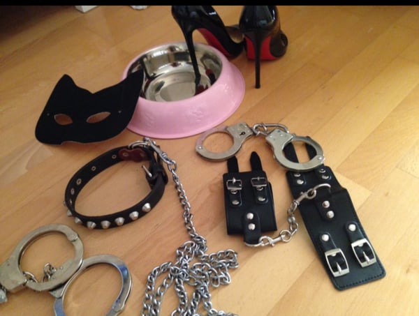 A collection of BDSM-themed accessories including handcuffs, collars, high heels, a mask, and a bowl arranged on a wooden floor.