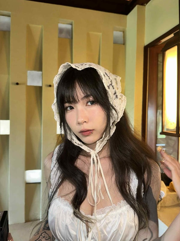 Young woman wearing a lace bonnet and dress sitting indoors with soft lighting and wooden architecture in the background.
