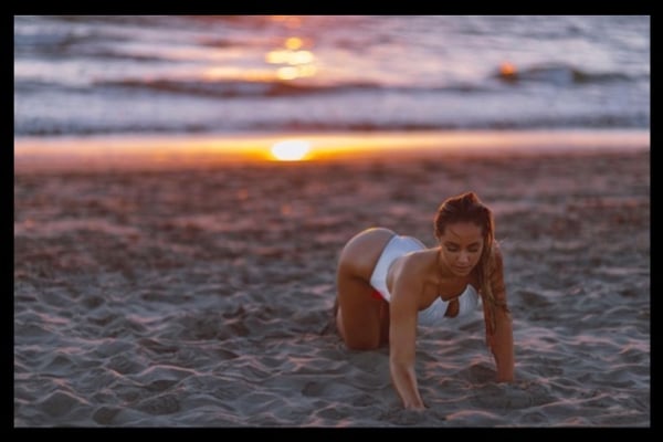 A woman in a white swimsuit posing on a sandy beach at sunset with the ocean in the background.