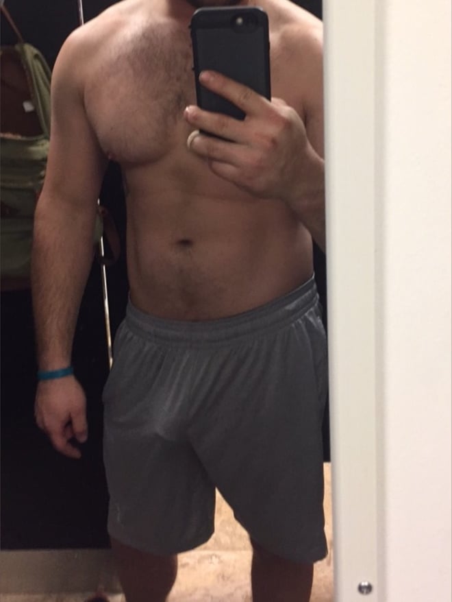 A muscular man taking a mirror selfie in athletic shorts, standing in a locker room with gym bags nearby.