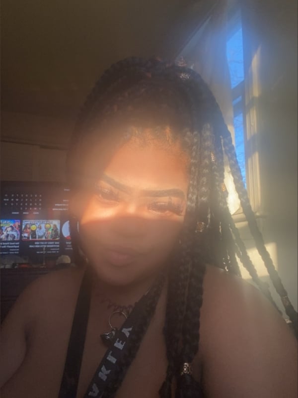 Young woman with long braided black hair posing topless in a sunlit room with natural light highlighting her face.