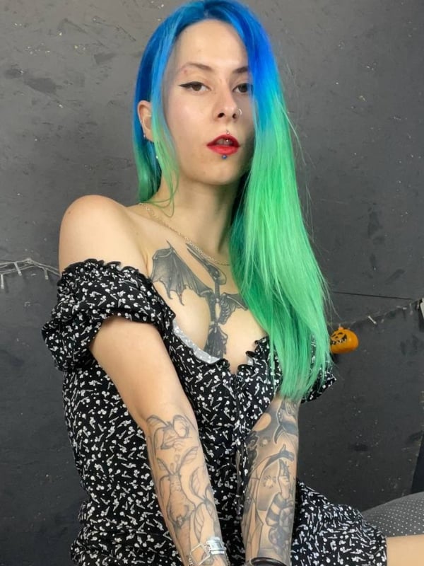 A woman with vibrant blue-green dyed hair and tattoos sits wearing an off-shoulder top in a room with Halloween decorations.