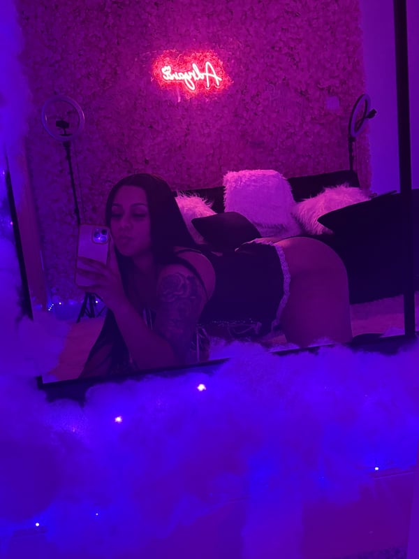 A woman with long dark hair poses on a fluffy white bed in a room decorated with pink floral walls, neon signage, and purple lighting.