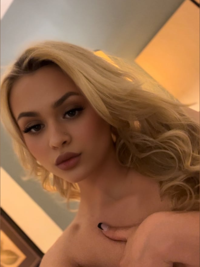 Close-up portrait of a young woman with long wavy blonde hair, subtle makeup, and hoop earrings, posing with her hand near her shoulder against a warm-toned background.