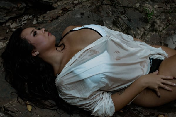A woman reclining on rocks in a natural outdoor setting, wearing a white sheer top and black bottoms, with wet hair and a relaxed pose.
