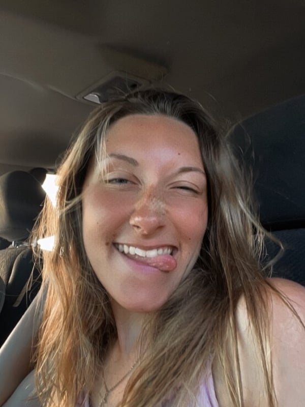 A smiling young woman with blonde hair and a nose piercing sticks out her tongue while sitting in a car, wearing a light purple top.