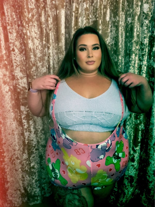 A woman with long brown hair stands in front of sparkly curtains, wearing a light blue top and colorful floral-patterned overalls.