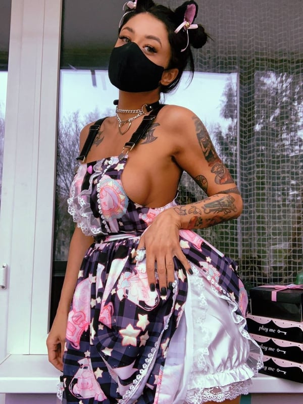 A woman standing indoors near a window, wearing a colorful outfit with cat ears and a face mask.