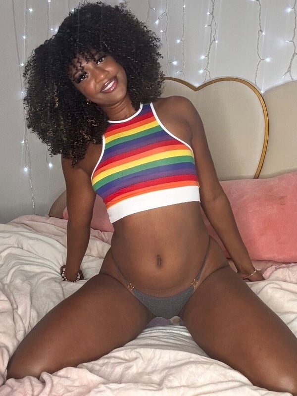Young woman with curly hair smiling on a bed, wearing a rainbow-colored halter top and bottoms, surrounded by pillows and string lights.