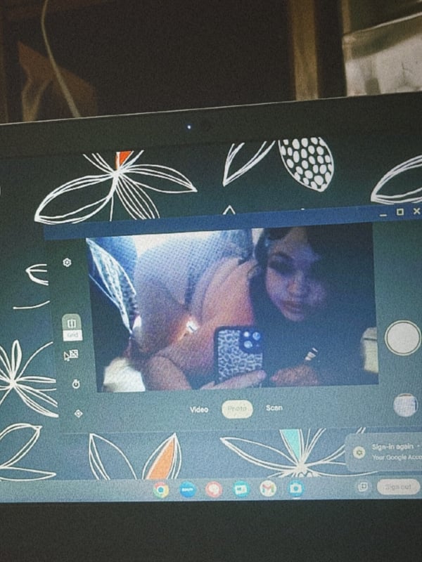 A laptop screen displaying a close-up selfie of a woman with dark hair in a dimly lit setting, surrounded by decorative leaf patterns on a greenish background.
